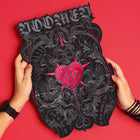 Two hands hold the Doomed - Custom Initials Embroidered Patch, a black shield-shaped piece with ornate silver designs, occult symbols, a stitched red heart, and bold WOMEN lettering, standing out against a vivid red background.