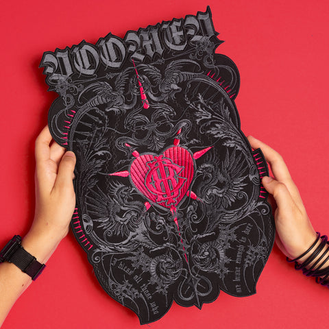 Two hands hold the Doomed - Custom Initials Embroidered Patch, a black shield-shaped piece with ornate silver designs, occult symbols, a stitched red heart, and bold WOMEN lettering, standing out against a vivid red background.