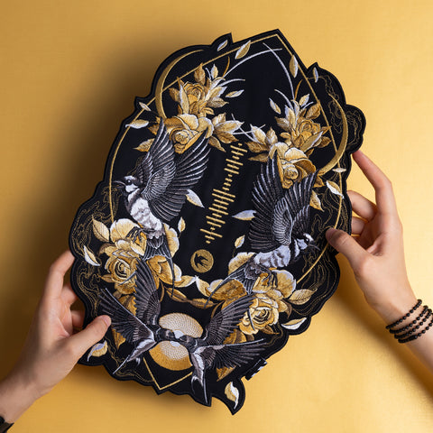 Two hands hold a Spotify Song Code Embroidered Patch featuring gold and white roses, two flying birds, and geometric designs on a dark background, placed against a yellow surface.