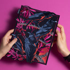 Two hands hold the Four Crows Embroidered Back Patch, showing four black crows perched on pink and red branches with leaves and berries, set against a vibrant pink background.