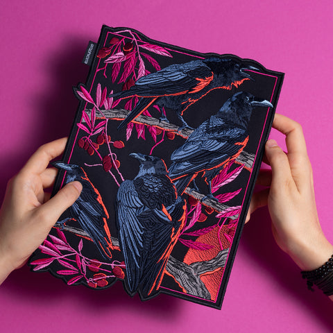 Two hands hold the Four Crows Embroidered Back Patch, showing four black crows perched on pink and red branches with leaves and berries, set against a vibrant pink background.