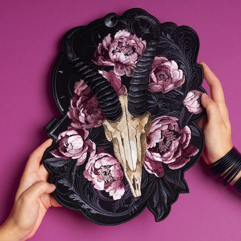 Two hands hold the Rose Ash Skull and Flowers Embroidered Patch, which features a horned animal skull with pink peonies on a black ornate background, set against a solid magenta surface.