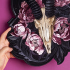 A hand holds the Rose Ash Skull and Flowers Embroidered Patch, featuring a goat skull with black horns and pink and purple peonies on a dark background, set against a solid magenta surface.