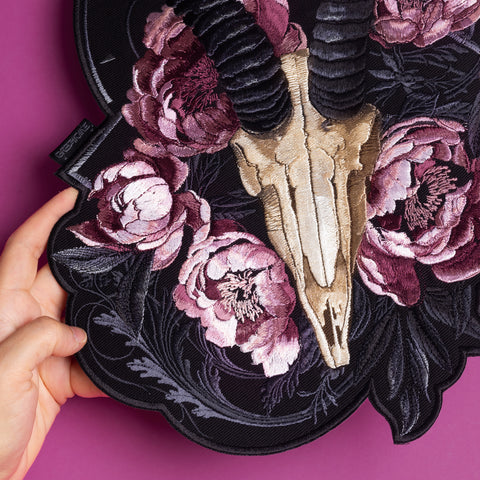A hand holds the Rose Ash Skull and Flowers Embroidered Patch, featuring a goat skull with black horns and pink and purple peonies on a dark background, set against a solid magenta surface.