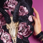 A hand holds the Rose Ash Skull and Flowers Embroidered Patch, which displays a gold animal skull with black horns and pink peonies on a black background, set against a magenta surface.