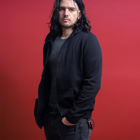 A person with long dark hair, wearing the Baphomet Unisex Embroidered Hoodie styled in gothic streetwear, stands against a solid red background with a neutral expression and one hand in their pocket.