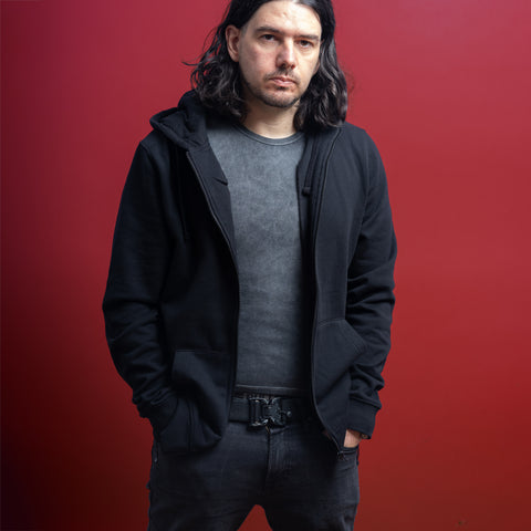 A man with long dark hair wears a black Baphomet Unisex Embroidered Hoodie over a gray tee, standing against a red background with his hands in his pockets, looking seriously at the camera and channeling a gothic streetwear vibe.