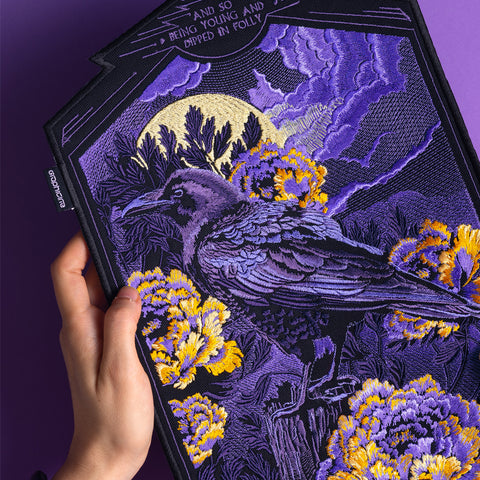 A hand holds the Crow and Peonies Embroidered Back Patch, showing a black crow among yellow peonies with a full moon and purple sky. The text above says, AND BEING YOUNG AND DIPPED IN FOLLY—a striking moonlit design.