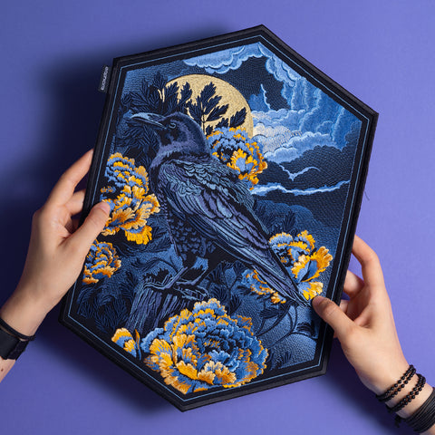A person holds the Crow & Peonies Embroidered Back Patch, a hexagonal patch featuring a gothic raven among yellow and blue peonies, dramatic clouds, and a full moon, all shown on a purple surface.