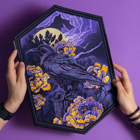 Two hands hold the Crow & Peonies Embroidered Back Patch, which features a gothic raven perched among yellow flowers, with a full moon and purple clouds in the background, displayed against a purple surface.