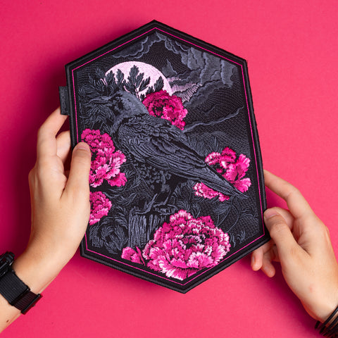 Two hands hold the Crow & Peonies Goth Embroidered Patch, which shows a black crow among pink flowers, a full moon, and dark clouds, all set against a bright pink background.