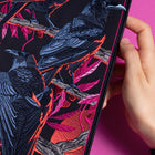 A hand displays the Four Crows Embroidered Back Patch, showing detailed black ravens on branches with pink and red leaves, set against a bright pink background.