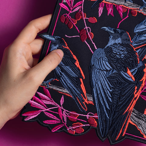 A hand holds the Four Crows Embroidered Back Patch, showing black fabric with mystical embroidery of four black crows on tree branches, surrounded by pink and red leaves and berries on a vibrant magenta background.
