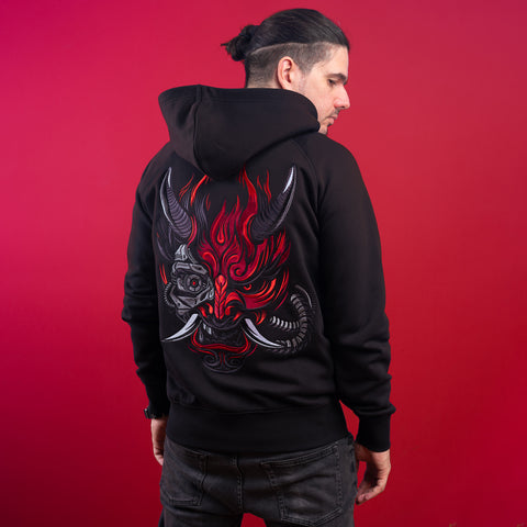 A man with dark hair in a bun models the Cyberpunk Oni Unisex Embroidered Hoodie, featuring a bold, colorful demon mask with red flames and horns on the back, set against a red background, epitomizing Japanese streetwear style.