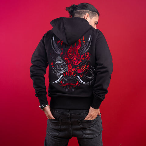 A person with long dark hair in a ponytail stands against a solid red background, hands in their jeans’ back pockets, wearing the Cyberpunk Oni Unisex Embroidered Hoodie featuring a bold red and gray oni mask design on the back.