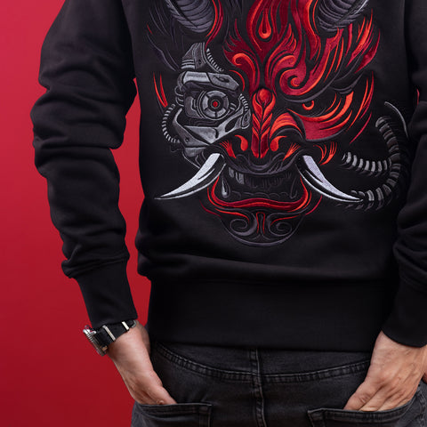 A person wearing the Cyberpunk Oni Unisex Embroidered Hoodie, featuring a bold, colorful Oni mask embroidered on the back, stands against a red background with hands in their jeans pockets—delivering edgy Japanese streetwear style.