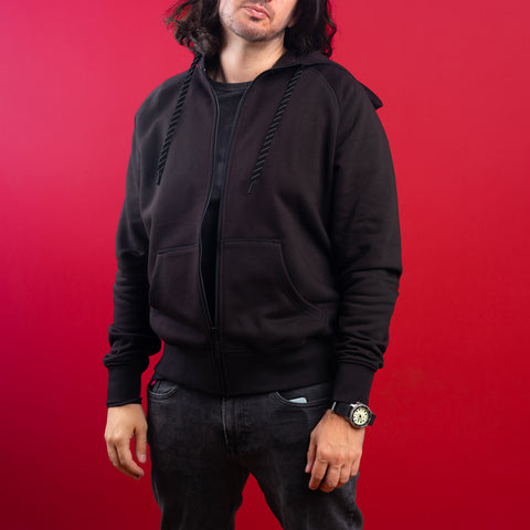 A person in a black Cyberpunk Oni Unisex Embroidered Hoodie and black jeans stands against a red background, one hand by their side and the other in a pocket. The hoodie has front pockets, drawstrings, and an embroidered Oni mask inspired by Japanese streetwear.