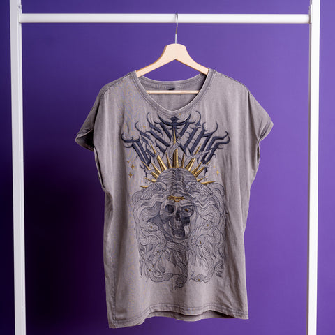 The Gorgone Embroidered Female T-shirt, featuring a gray acid-wash finish and a skull with flowing hair, golden crown, and bold text graphic, hangs on a wooden hanger against a purple background for an edgy look.