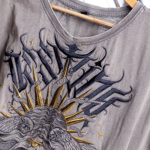 A close-up of the Gorgone Embroidered Female T-shirt in grey on a wooden hanger, featuring two snakes, gold sun rays, stars, and bold black lettering—crafted with unique garment dye for standout style and character.