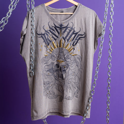 The Gorgone Embroidered Female T-shirt features a gray acid-wash design with a detailed skull-faced figure, gold ray headpiece, and gothic motifs, displayed on a wooden hanger against a purple backdrop with metal chains.