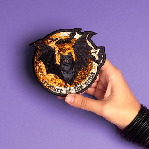 A hand holds the Bats Halloween Embroidered Patch, showing a black bat with spread wings on an orange background and a "guardian of the night" banner, resting on a purple surface. Ideal as an iron-on Halloween accessory.