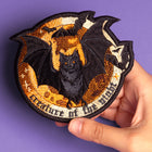 A hand holds the Bats Halloween Embroidered Patch, showing a black bat with outspread wings in front of an orange moon, surrounded by stars and smaller bats, all displayed on a purple surface.