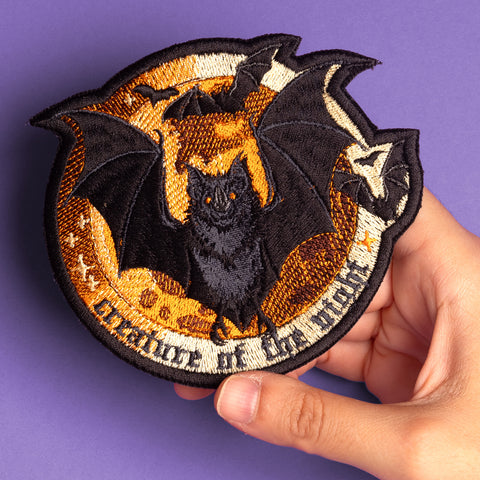 A hand holds the Bats Halloween Embroidered Patch, showing a black bat with outspread wings in front of an orange moon, surrounded by stars and smaller bats, all displayed on a purple surface.