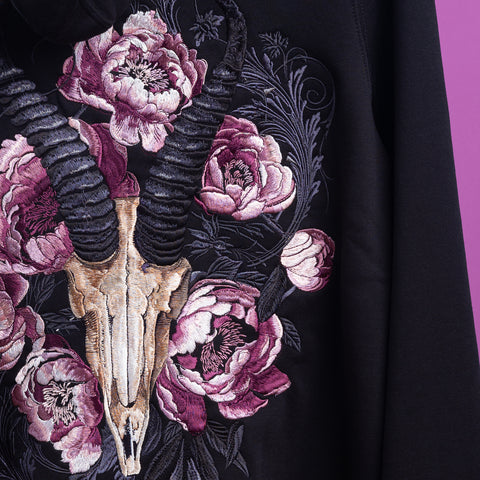 The Blesbok Unisex Embroidered Hoodie showcases a ram skull with curved horns, surrounded by pink and purple peonies and leafy accents on a purple backdrop for a bold dark pastel goth look.
