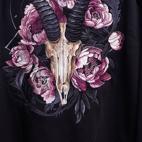 The Blesbok Unisex Embroidered Hoodie features a bold ram skull with curved black horns, surrounded by pink and purple peonies—ideal for fans of cozy, unique pastel goth style and dark pastel aesthetics.