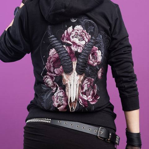 Wearing the Blesbok Unisex Embroidered Hoodie featuring a ram skull and pink flowers on the back, a figure stands against a purple background with studded belt and wristbands, embodying pastel goth style while facing away from the camera.