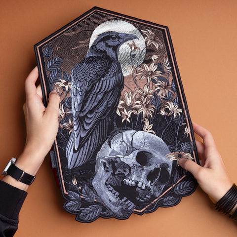 A person holds the Raven and Skull Embroidered Back Patch, featuring a dark raven perched on a skull among lilies and a full moon, capturing the mysterious allure of gothic embroidered patches.