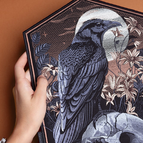 A hand holds the Raven and Skull Embroidered Back Patch, featuring a black raven on a skull with a full moon and lilies on hexagonal fabric—ideal for gothic fashion accessories.
