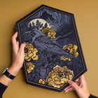 Two hands hold the Crow & Peonies Goth Embroidered Patch, showcasing a black crow on a stump among yellow flowers beneath a full moon and clouds—ideal for adding Victorian-inspired gothic style to your outfit.