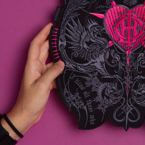 A hand displays the Doomed - Custom Initials Embroidered Patch in black and pink, featuring intricate heart, dragon, and occult designs. Set against a bold magenta background, it captures a striking alternative fashion aesthetic.