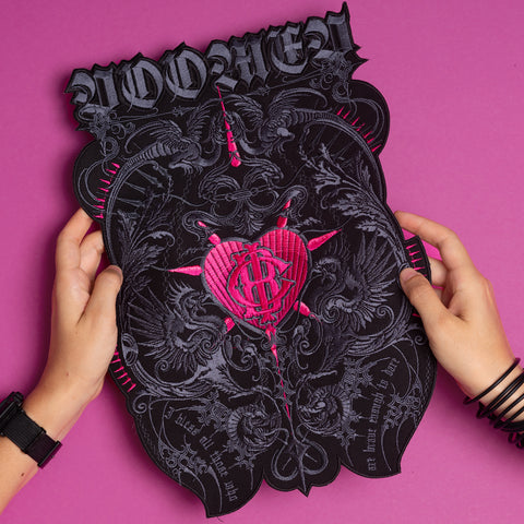 Two hands hold the Doomed - Custom Initials Embroidered Patch, a black and pink design with ornate patterns, a heart featuring a dollar sign at its center, and "WOMEN" in bold lettering, set against a vibrant pink background.