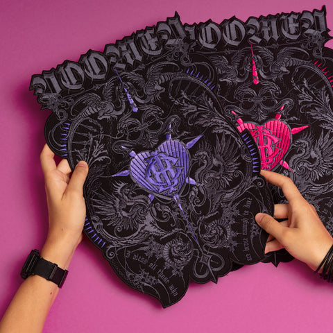 Two hands hold three intricate black cut-out signs on a pink surface, each with ornate patterns and a Doomed - Custom Initials Embroidered Patch in purple, blue, or pink, blending occult symbolism with decorative details.