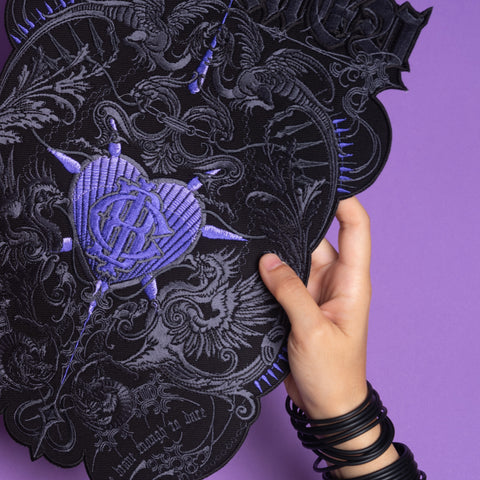 A hand holds the "Doomed - Custom Initials Embroidered Patch," a black and purple piece with heart, sword, and occult motifs. The wearer’s black bracelets add alternative style, all set against a purple background.