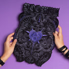 Two hands hold the Doomed - Custom Initials Embroidered Patch, featuring ornate black and purple embroidery, intricate designs, gothic lettering, and occult motifs—a bold, alternative fashion accessory shown on a purple background.