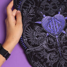 A hand holds an ornate black fabric with purple stitching, featuring the "Doomed - Custom Initials Embroidered Patch" with a heart, wings, and intricate patterns on a purple background. The person wears a black wristwatch.