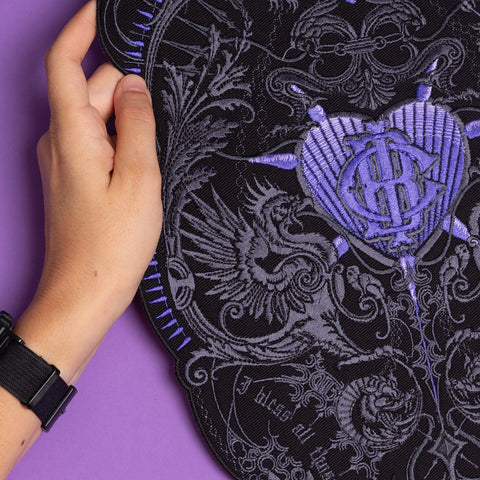A hand holds an ornate black fabric with purple stitching, featuring the "Doomed - Custom Initials Embroidered Patch" with a heart, wings, and intricate patterns on a purple background. The person wears a black wristwatch.