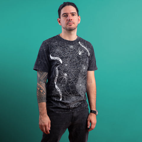 A man with long dark hair tied back wears the Spider Web Embroidered Glow In The Dark Unisex T-shirt, featuring a white spider web on black, against a teal background—an eye-catching, unique Halloween gift. He has a tattoo and a black watch.