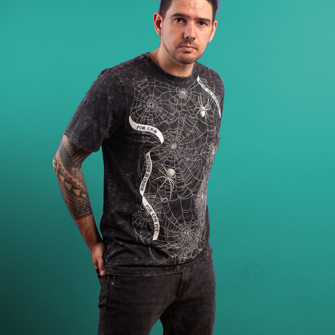 A tattooed man stands against a teal background, hands in pockets, wearing the Spider Web Embroidered Glow In The Dark Unisex T-shirt with white spiderweb graphics, looking slightly to the right.