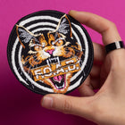 A hand holds the Hallow Kitty Embroidered Patch, showing a fierce multicolored cat with an open mouth over black-and-white spiral stripes, “ROAD” across its mouth, and a bright pink background.