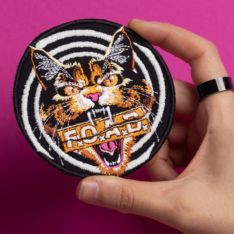 A hand holds the Hallow Kitty Embroidered Patch, showing a fierce multicolored cat with an open mouth over black-and-white spiral stripes, “ROAD” across its mouth, and a bright pink background.