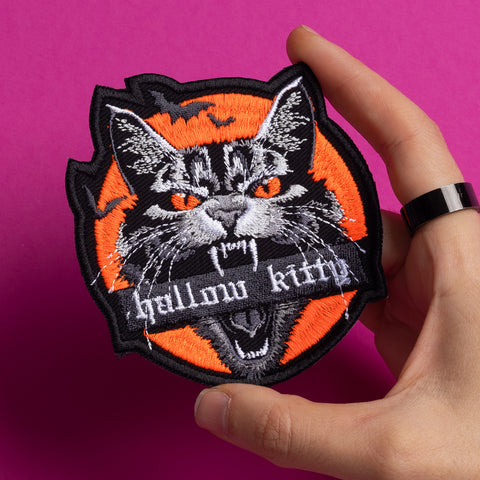 A hand holds the Hallow Kitty Embroidered patch, showing a fierce black cat with orange eyes and fangs on an orange circle, “Hallow kitty” stitched on its mouth, set against a bright pink background.