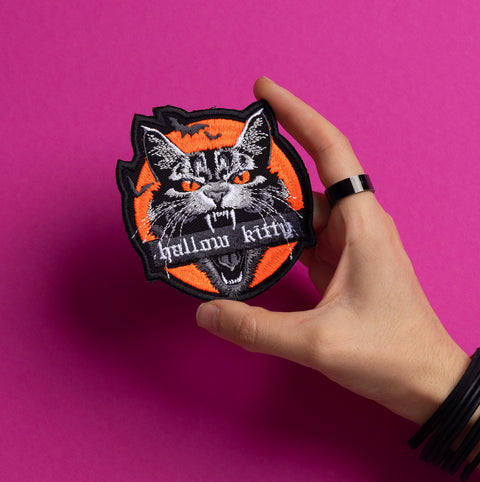 A hand holds the Hallow Kitty Embroidered Patch—a fierce, fanged black cat on an orange background with "hallow kitty" text. This patch pops against a pink backdrop and is perfect for adding a spooky Halloween touch!.