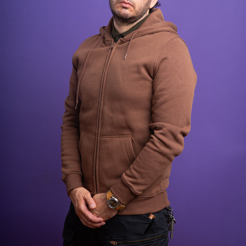 A person wears a brown Skull Unisex Embroidered Hoodie and black pants, standing against a solid purple background with hands loosely clasped in front, highlighting a modern gothic streetwear vibe.