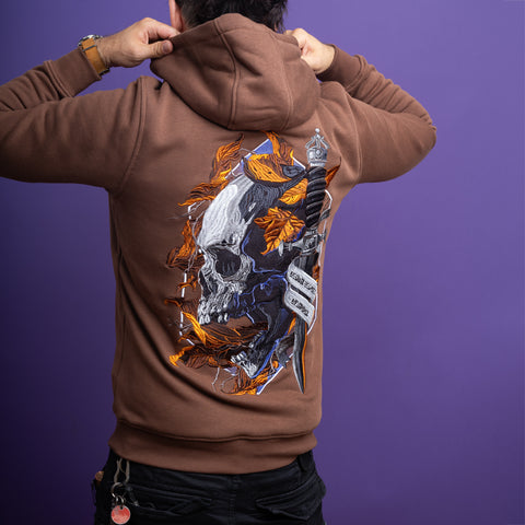A person in a brown Skull Unisex Embroidered Hoodie, featuring a bold skull, dagger, and orange leaves design, faces away adjusting their hood against a purple background. Keys hang from their belt, emphasizing the gothic streetwear vibe.