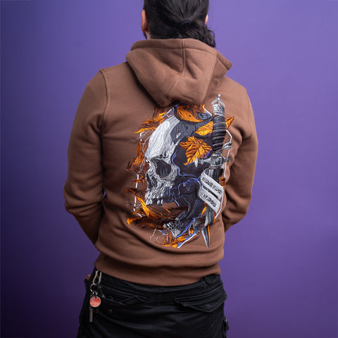 A person with long hair in a ponytail faces away, wearing the Skull Unisex Embroidered Hoodie in brown with a large skull, autumn leaves, and dagger design. Keys hang from their pocket against a purple background.