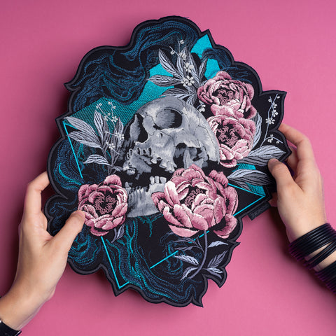 A person holds the Skull and Flowers Embroidered Back Patch, featuring a skull with pink flowers and teal leaves on a dark, ornate background atop a pink surface—ideal for gothic fashion enthusiasts.